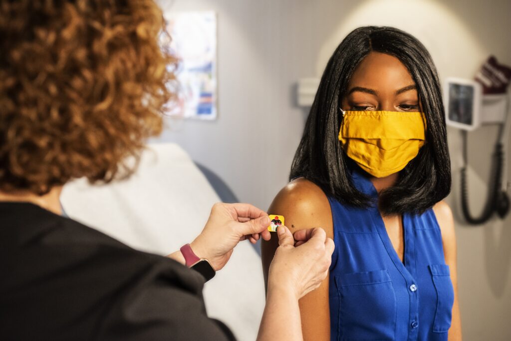 A Black woman with a mask on getting a bandaid after receiving a vaccination | To Get Vax or not?